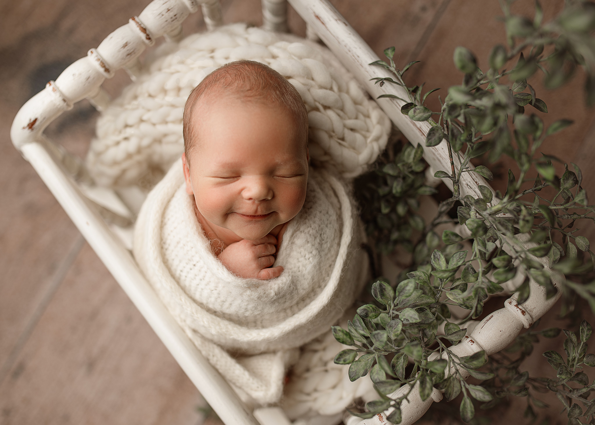 baby boy smiling for his newborn photoshoot in a white basket prop with greenery wrapped in a white wrap for a natural look